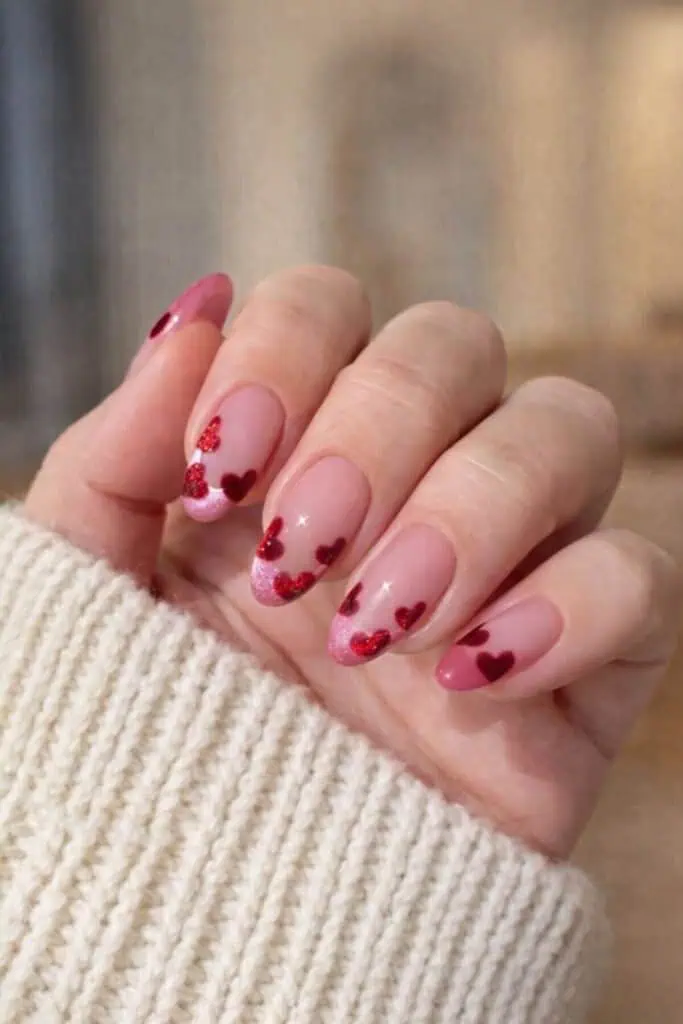 Close-up of woman's hand showing almond-shaped nails with soft pink base, subtle pink glitter French tips, and scattered small red glitter hearts on each nail, held against a cozy sweater sleeve in warm indoor light.