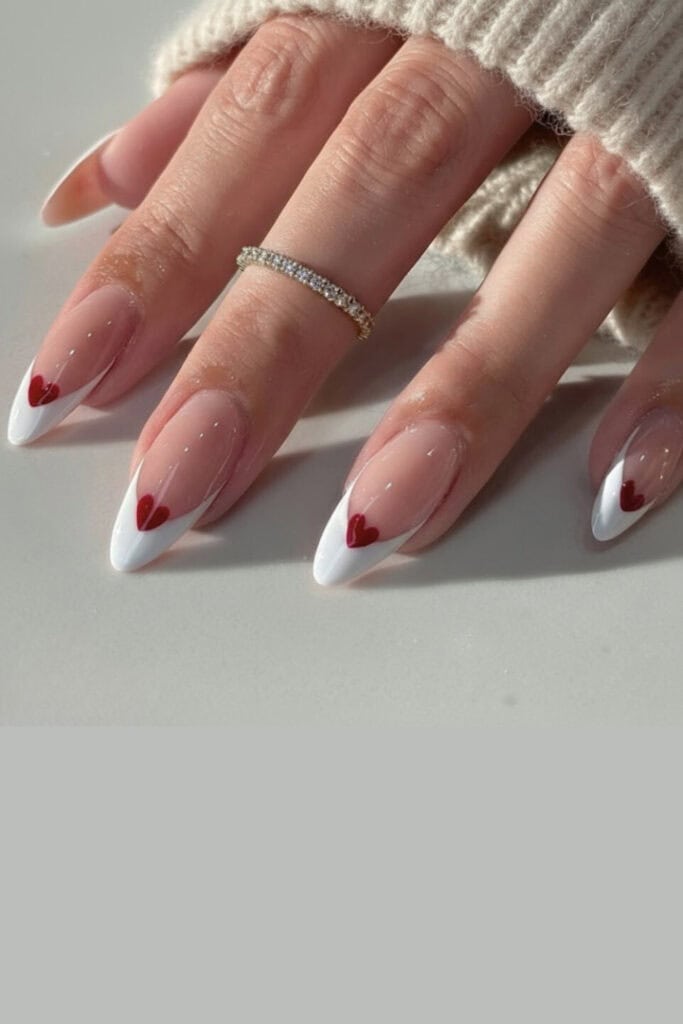 Close-up of woman's hand with long almond-shaped nails featuring nude-pink base, crisp white French tips, small red heart accent on each nail, glossy finish, subtle sparkle, held against beige knit sweater sleeve in warm indoor light.