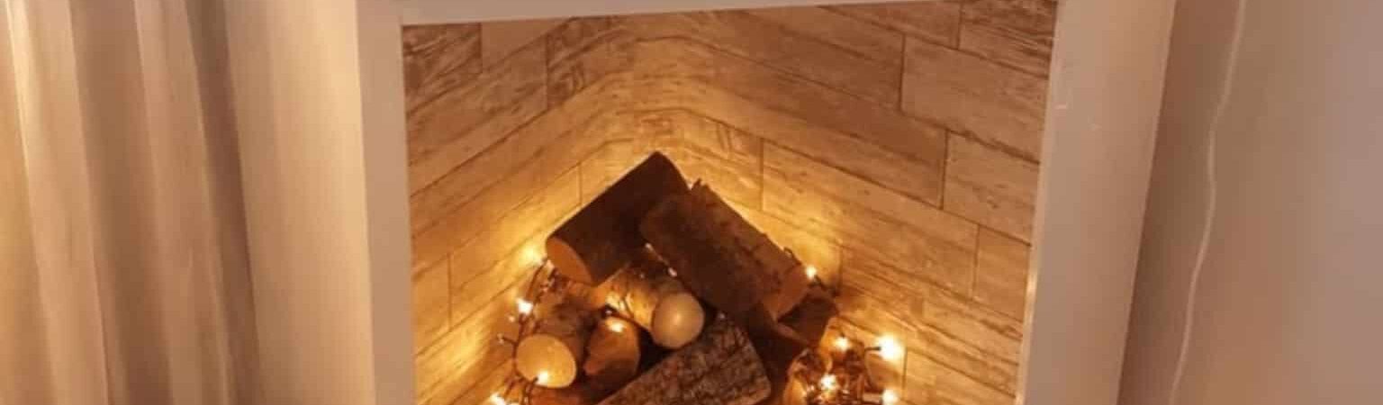 White faux fireplace in a cozy apartment decorated with stacked logs, warm string lights, candles, and neutral fall decor.