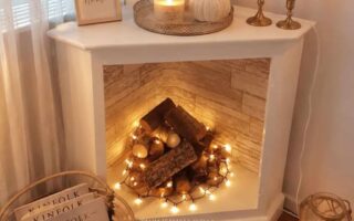 White faux fireplace in a cozy apartment decorated with stacked logs, warm string lights, candles, and neutral fall decor.