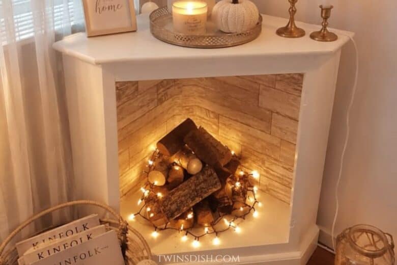 White faux fireplace in a cozy apartment decorated with stacked logs, warm string lights, candles, and neutral fall decor.