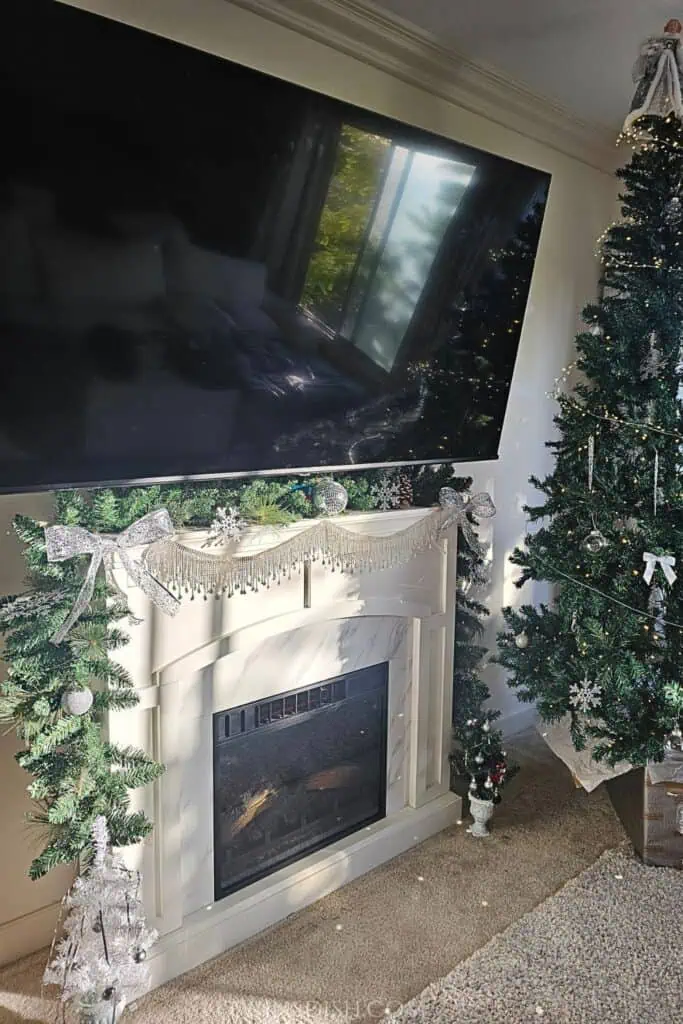 Christmas living room with an electric fireplace, decorated mantel garland, silver bows, a large TV, and a Christmas tree with white lights.