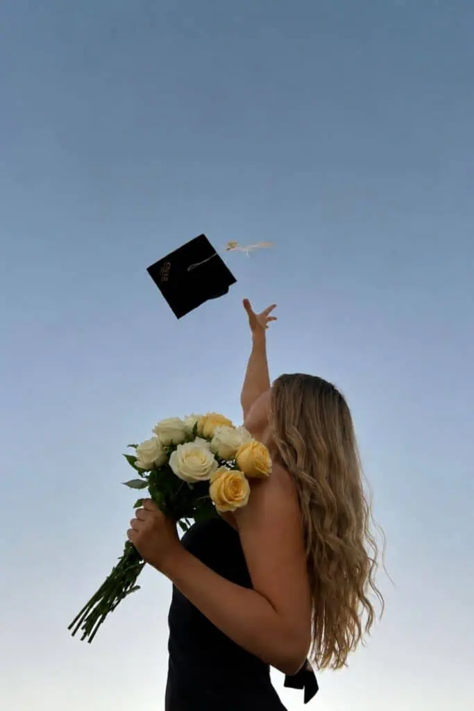 Girl in black dress tossing graduation cap while holding white and yellow roses against blue sky, creative graduation picture ideas pose for a photoshoot.