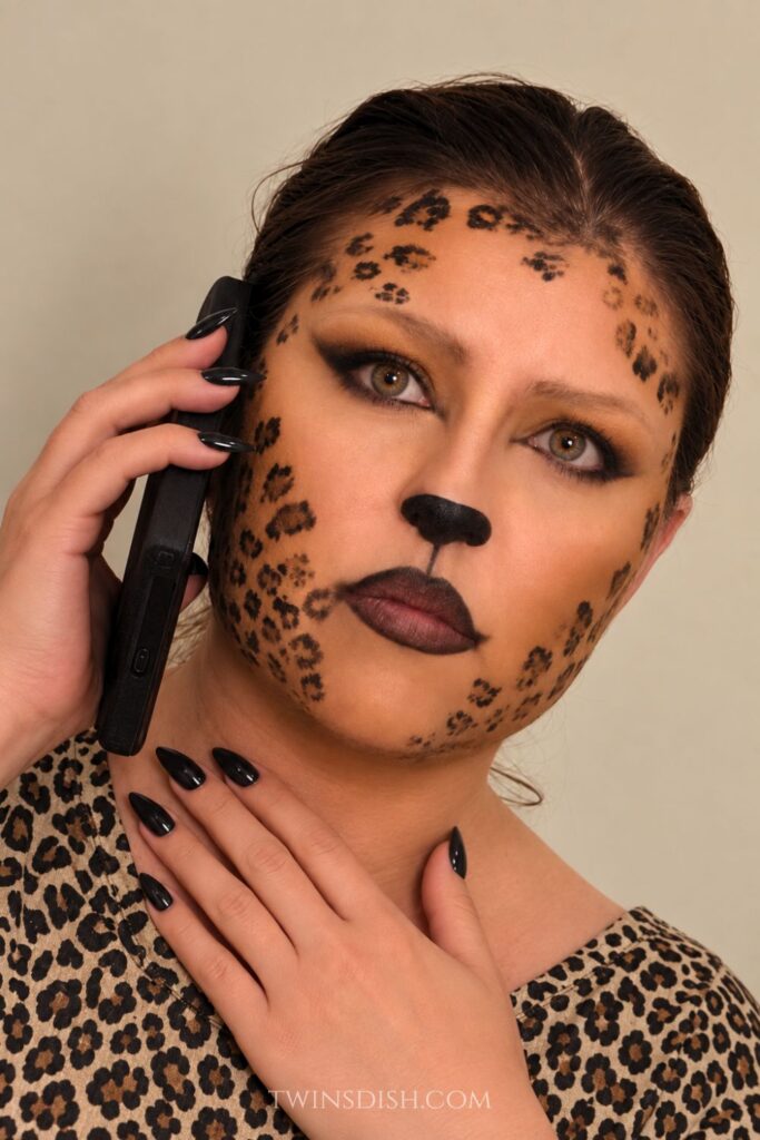 Woman wearing cheetah Halloween makeup with leopard spots, smoky eye makeup, black nose makeup, long black nails, and a leopard print top.