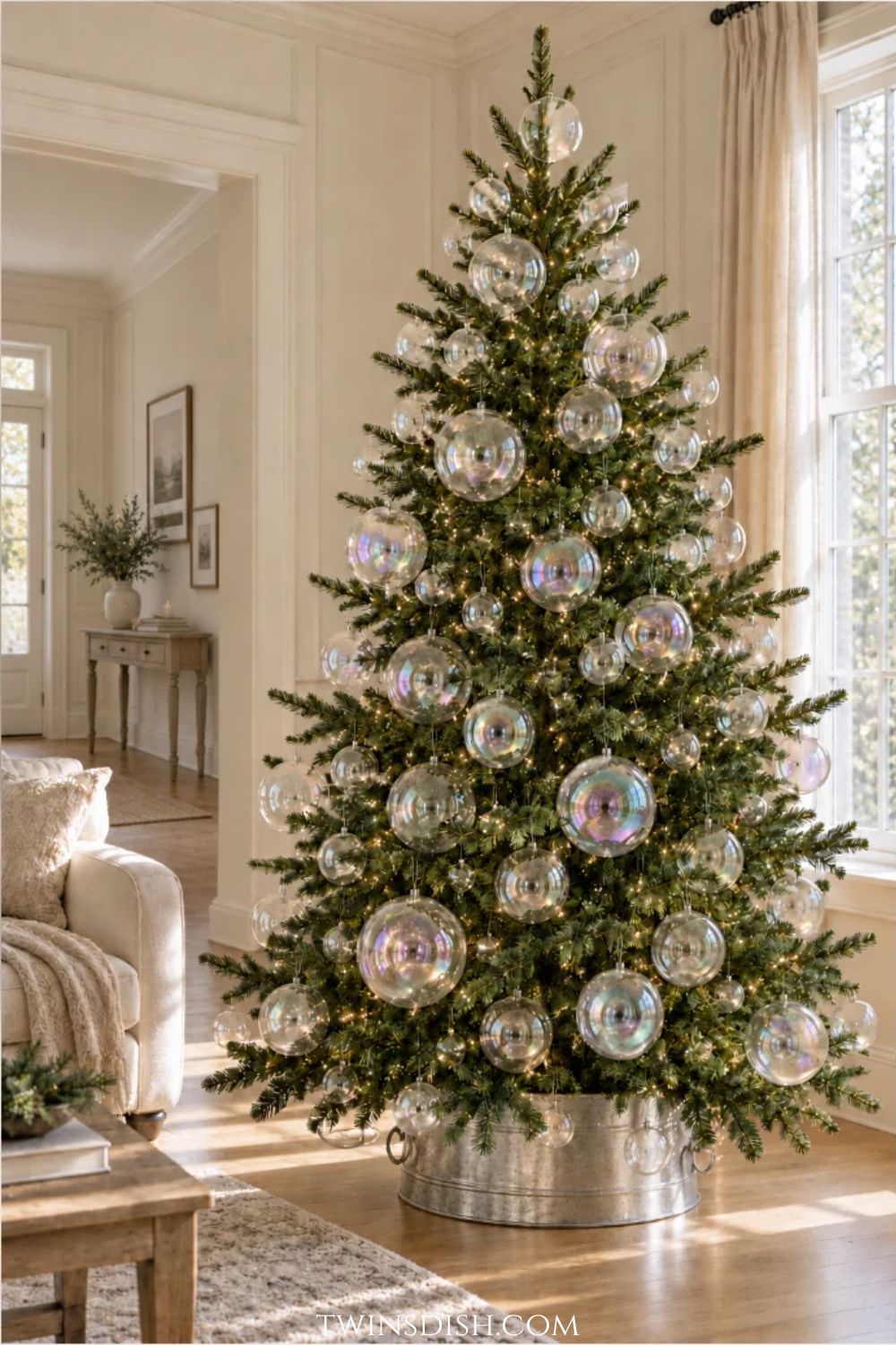 Full-length Christmas tree decorated with clear and iridescent soap bubble ornaments, warm white twinkle lights, and a silver metal tree collar beside a sunny living room window.