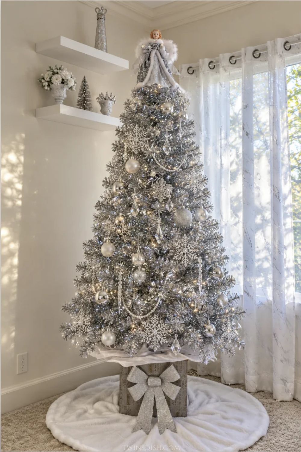 Silver metallic tinsel Christmas tree decorated with crystal snowflakes, silver and pearl ornaments, hanging icicles, white lights, and an angel topper, displayed on a raised tree stand with a large glitter bow.