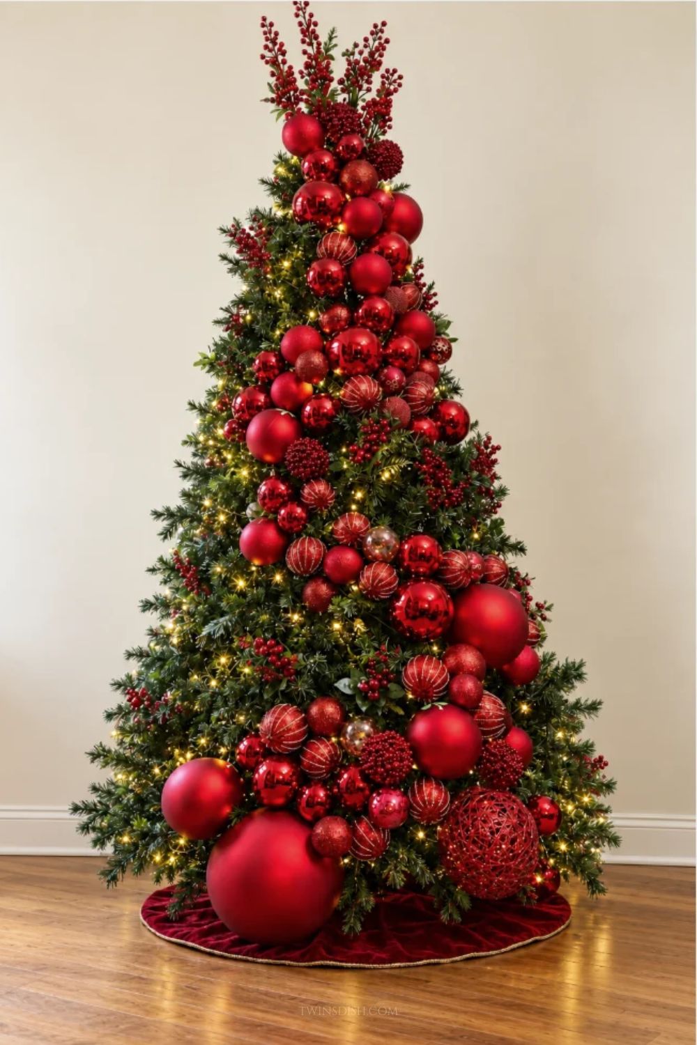 Elegant red Christmas tree decorated with oversized red ornaments, glossy and matte baubles, glittered ornaments, red berry sprays, and warm white lights against a simple cream wall.