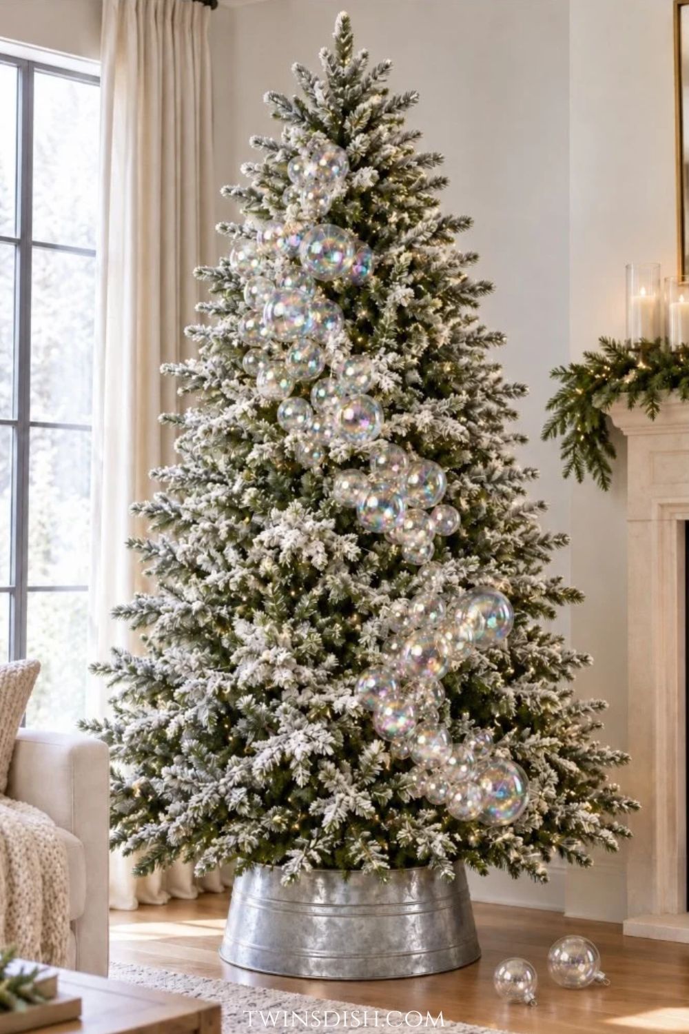 Full-length flocked Christmas tree decorated with cascading clusters of clear iridescent bubble ornaments and warm white lights, displayed in a silver metal tree collar with two glass ornaments resting on the floor.